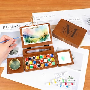 Op de afbeelding: Een houten aquarelverfset met een scharnierend deksel, een ingebouwd palet van 24 kleuren en een aquarelkwast. De set bevat een kleine aquarelverfschildering op een klembord en een kleine goudkleurige kom. De set staat op een wit oppervlak.