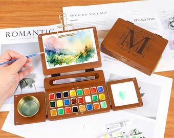 May include: A wooden watercolor paint set with a hinged lid, a built-in palette of 24 colors, and a watercolor brush. The set includes a small watercolor painting on a clip board and a small gold-colored bowl. The set is on a white surface.