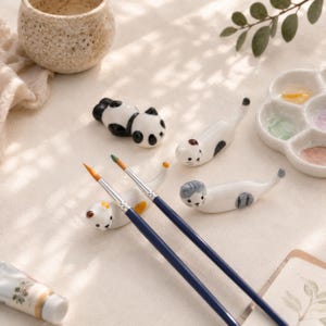 Könnte beinhalten: Ein Set aus vier keramikfarbenen Pinselablagen in Tierform, darunter ein Panda und drei Katzen, zwei dunkelblaue Pinsel mit goldfarbenen Borsten, eine weiße Keramik-Farbpalette mit Farbresten und ein kleiner, gesprenkelter Keramikbecher.