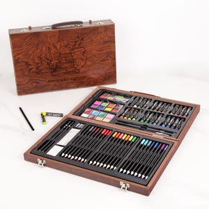 Könnte beinhalten: Ein Holzkoffer mit Kunstbedarf. Der offene Koffer zeigt Farbstifte, Aquarellfarben, Ölpastelle und Pinsel. Der geschlossene Koffer hat einen Griff und ein geätztes Design mit dem Text "Annie's art supplies."