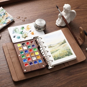 May include: A watercolor painting set with a brown leather case. The set includes a wooden palette with various colors, a white palette with mixed paints, a small metal container, a paintbrush, and a small watercolor painting of a landscape. A small white angel statue is also present.