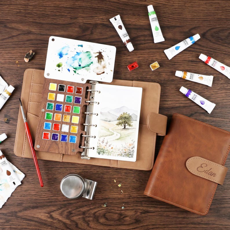 May include: A brown leather journal with a watercolor palette, paint tubes, and a watercolor painting of a landscape. The palette has a variety of colors, and the journal is open to a blank page. A small metal clip is also visible.