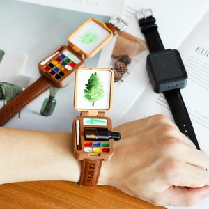 May include: Brown leather strap watch with a miniature artist's palette design. The watch face opens to reveal a tiny painting of a tree. The palette includes various colours of paint and a small brush. The watch strap is embossed with the letters "M&D".