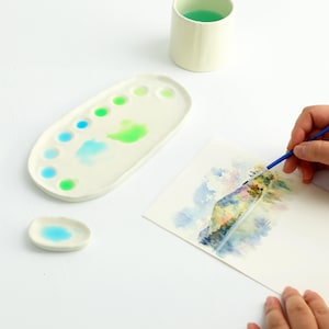 Könnte beinhalten: Eine weiße Keramik-Aquarellpalette mit blauer und grüner Farbe. Eine kleine, ovale Palette mit blauer Farbe befindet sich darunter. Eine Person malt mit einem blaugriffigen Pinsel auf Aquarellpapier. Eine weiße Keramiktasse mit grüner Flüssigkeit befindet sich im Hintergrund.