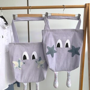 Op de afbeelding: Twee lavendelkleurige corduroy tote tassen met cartoongezichten, steraccenten en bungelende benen. Elke tas heeft zwarte ogen, witte oogleden en stervormige patches. De benen zijn voorzien van een blauw-wit geruit patroon.