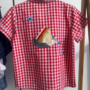 Könnte beinhalten: Kurzärmeliges Hemd mit rot-weißem Vichy-Muster. Auf der Rückseite des Hemdes befindet sich ein Aufnäher mit einem Hut mit farbigem Streifen und zwei blauen Knöpfen. Das Hemd hängt an einem Holzbügel.