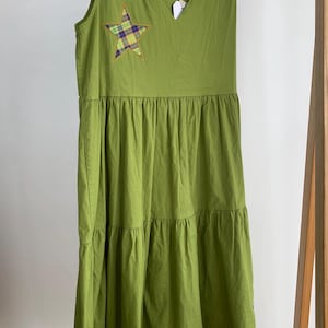 Może przedstawiać: Bezrękawnikowa, oliwkowa sukienka z dekoltem w serek i warstwową spódnicą. Aplikacja w kształcie gwiazdy w kratę jest przyszyta do klatki piersiowej. Sukienka wisi na drewnianym wieszaku.