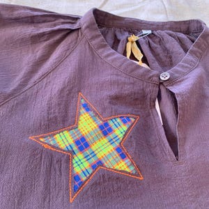 May include: A plum-colored top with a mandarin collar and a star-shaped patch. The star is plaid with neon green, blue, and orange accents. The top has a keyhole neckline and a single button closure.