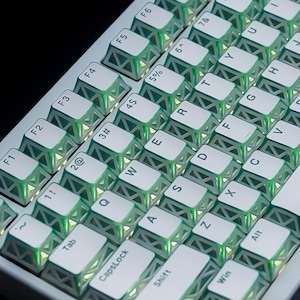 May include: A computer keyboard featuring white keycaps with a structural, open-frame lattice design on the sides. The keys are illuminated from beneath with a bright green light, highlighting the intricate geometric patterns of the keycaps.