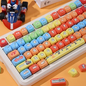 Pode incluir: Um teclado mecânico branco com teclas coloridas exibindo letras, números e gráficos com o tema Super Mario. A barra de espaço apresenta um design de estrada e uma pequena figura de Mario kart está posicionada ao lado do teclado.