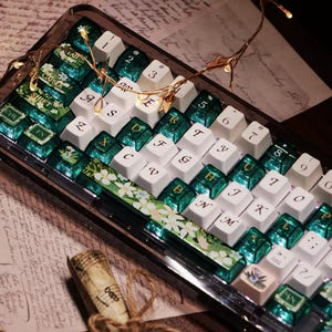 May include: A custom mechanical keyboard with white and green keycaps. The keycaps feature floral designs and transparent green accents. The keyboard has a clear case and is illuminated with small fairy lights.