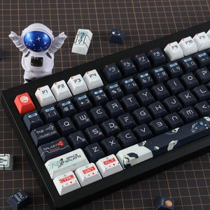 May include: A black mechanical keyboard with white and dark blue keycaps. The keycaps feature space-themed designs and text. An astronaut figurine is to the left. Additional keycaps are scattered around.