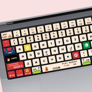 Peut inclure: Une protection de clavier d'ordinateur portable au thème de jeu vidéo rétro, avec des touches couleur crème, des lettres pixelisées noires et des accents colorés. Le design inclut des graphismes de champignons, de personnages et de paysages, avec le texte 1 PLAYER GAME, 2 PLAYER GAME, SHIFT, PLAY AGAIN, YES et NO.