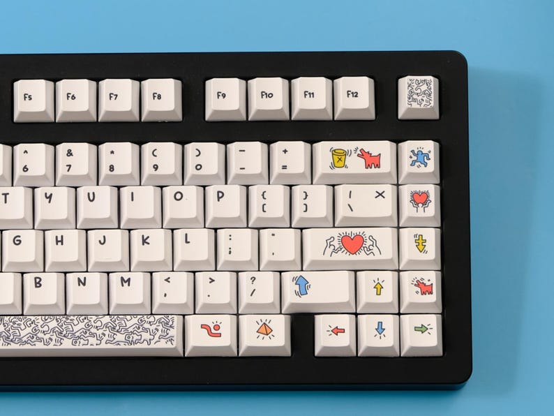 May include: A black mechanical keyboard with white keycaps. Some keycaps feature colorful Keith Haring-style artwork. The keyboard has a standard QWERTY layout with function keys and a blue background.