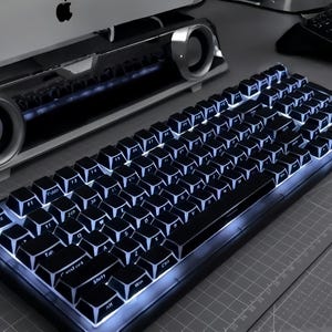 May include: A black mechanical keyboard with white illuminated keys. The keyboard is on a grey surface, with a computer monitor and speakers in the background. The keys are clearly labelled with white lettering.