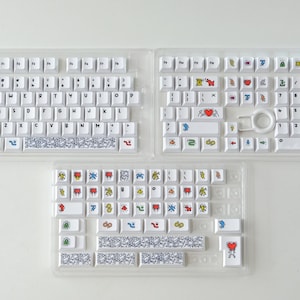May include: Three white keyboard sets with colorful graphic keycaps. The keys feature various designs, including hearts, figures, and abstract patterns. The keyboards are displayed in clear plastic trays, ready for assembly.