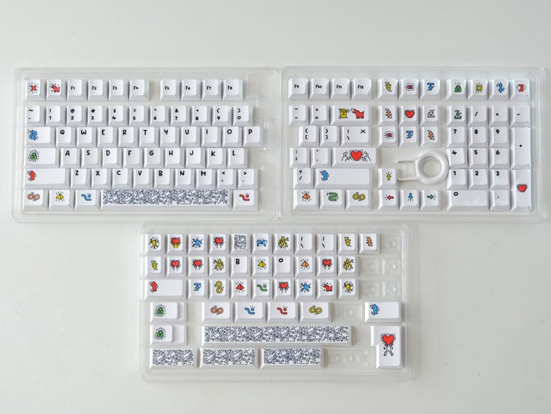 May include: Three white keyboard sets with colorful graphic keycaps. The keys feature various designs, including hearts, figures, and abstract patterns. The keyboards are displayed in clear plastic trays, ready for assembly.
