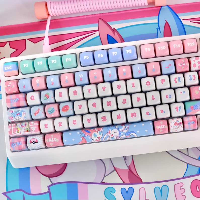 May include: A white mechanical keyboard with pastel pink, blue, and mint green keycaps. The keycaps feature various designs, including cartoon characters and food items. The keyboard rests on a matching pink and blue mousepad with a character design.