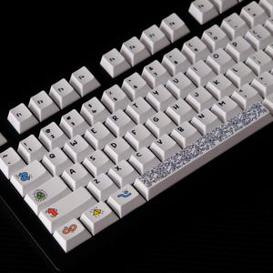 May include: White mechanical keyboard with custom keycaps. The keycaps feature various colorful icons and symbols, including hearts, arrows, and geometric shapes. A blue and white patterned space bar adds a decorative element.