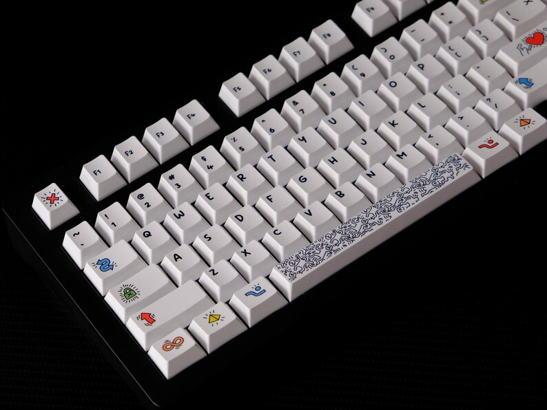 May include: White mechanical keyboard with custom keycaps. The keycaps feature various colorful icons and symbols, including hearts, arrows, and geometric shapes. A blue and white patterned space bar adds a decorative element.