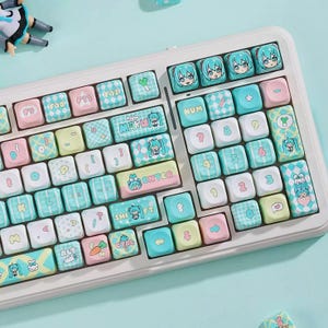 May include: A white keyboard with custom keycaps featuring pastel colors and anime-style character designs. The keycaps have pink, blue, and yellow accents, with various symbols and text. The keyboard is on a light blue surface.