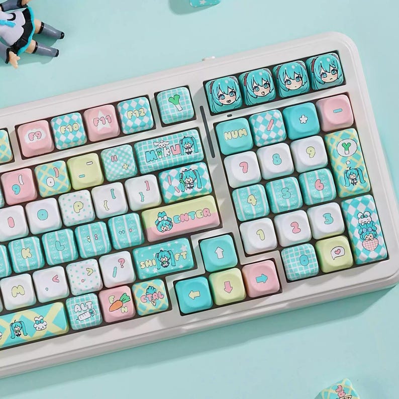 May include: A white keyboard with custom keycaps featuring pastel colors and anime-style character designs. The keycaps have pink, blue, and yellow accents, with various symbols and text. The keyboard is on a light blue surface.