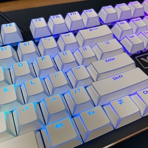 Op de afbeelding: Close-up van een wit mechanisch toetsenbord met blauwe LED-achtergrondverlichting. De toetsen zijn wit met zwarte letters. Het toetsenbord heeft een standaardindeling met functietoetsen, numerieke toetsen en pijltjestoetsen.
