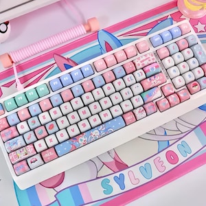 May include: A white mechanical keyboard with pastel pink, blue, and green keycaps. The keycaps feature various designs, including cartoon characters and patterns. A matching pink wrist rest and a mousepad with a similar design are also visible.