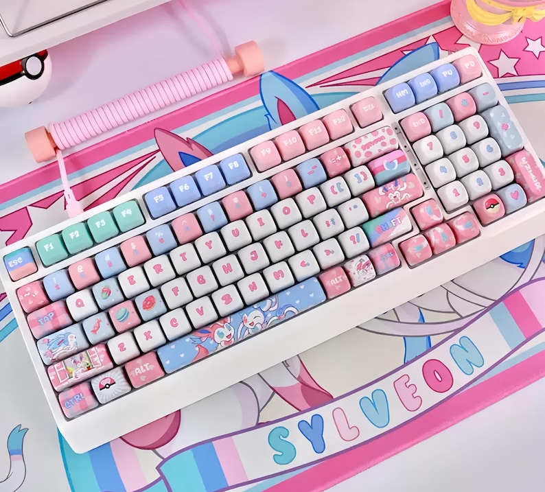 May include: A white mechanical keyboard with pastel pink, blue, and green keycaps. The keycaps feature various designs, including cartoon characters and patterns. A matching pink wrist rest and a mousepad with a similar design are also visible.