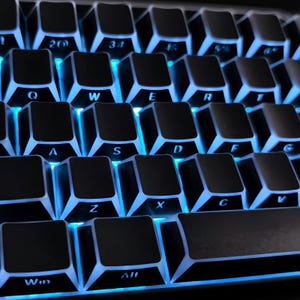 May include: A close-up shot of a black mechanical keyboard with blue illumination. The keys are black with white lettering, including letters, numbers, and symbols. The keyboard is angled, showing the key layout.