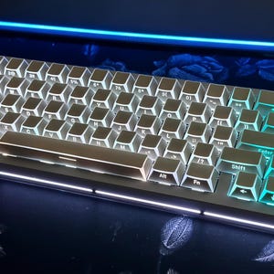 May include: A white mechanical keyboard with illuminated keys. The keyboard has a silver frame and teal-colored backlighting on the keys. The keys are labeled with white lettering. The keyboard is on a dark surface.
