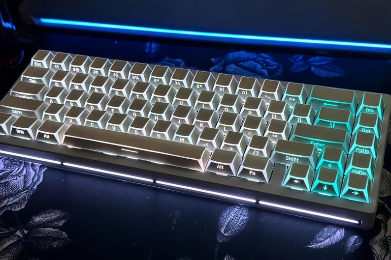 May include: A white mechanical keyboard with illuminated keys. The keyboard has a silver frame and teal-colored backlighting on the keys. The keys are labeled with white lettering. The keyboard is on a dark surface.