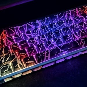 May include: A close-up of a mechanical keyboard with illuminated keys. The keys are black with a white cracked pattern, and the lighting transitions from red to purple to blue. The keyboard has a compact layout.