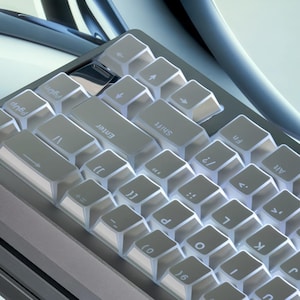 Op de afbeelding: Close-up van een grijs mechanisch toetsenbord met transparante keycaps. Het toetsenbord heeft een compacte indeling met verschillende symbolen en letters zichtbaar op de toetsen. De toetsen zijn verlicht, wat een heldere, moderne uitstraling cre&euml;ert.