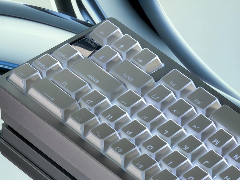 Op de afbeelding: Close-up van een grijs mechanisch toetsenbord met transparante keycaps. Het toetsenbord heeft een compacte indeling met verschillende symbolen en letters zichtbaar op de toetsen. De toetsen zijn verlicht, wat een heldere, moderne uitstraling cre&euml;ert.