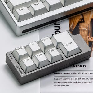 Op de afbeelding: Een close-up van een grijs en wit mechanisch toetsenbord met een apart numeriek toetsenbord. Het numerieke toetsenbord heeft witte toetsen met grijze letters. Het toetsenbord ligt op een wit oppervlak met bedrukt papier op de achtergrond.