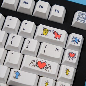 May include: A close-up of a mechanical keyboard with white keycaps. Some keys feature colorful Keith Haring-style artwork, including a red heart, a dog, and a figure. The keyboard has a black frame and is on a blue surface.
