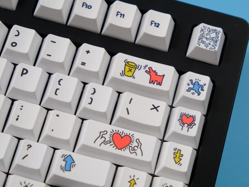 May include: A close-up of a mechanical keyboard with white keycaps. Some keys feature colorful Keith Haring-style artwork, including a red heart, a dog, and a figure. The keyboard has a black frame and is on a blue surface.