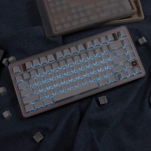 May include: A mechanical keyboard with white keycaps and blue backlighting. The keyboard has a metallic frame with a brown accent key. Several loose keycaps are scattered around the keyboard on a dark blue surface.