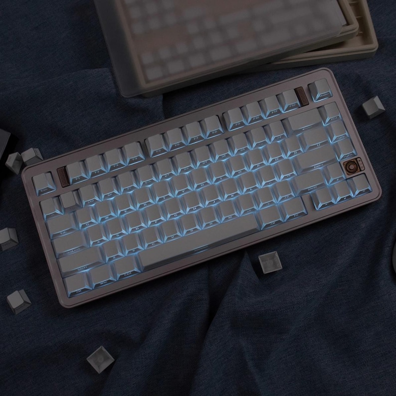 May include: A mechanical keyboard with white keycaps and blue backlighting. The keyboard has a metallic frame with a brown accent key. Several loose keycaps are scattered around the keyboard on a dark blue surface.