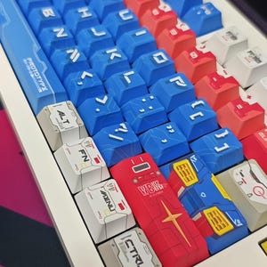 Puede incluir: Primer plano de un teclado mec&aacute;nico con teclas azules, rojas y blancas. Las teclas muestran letras, s&iacute;mbolos y texto japon&eacute;s. El teclado tiene un marco blanco y un lado azul con la palabra "PROTOTYPE".