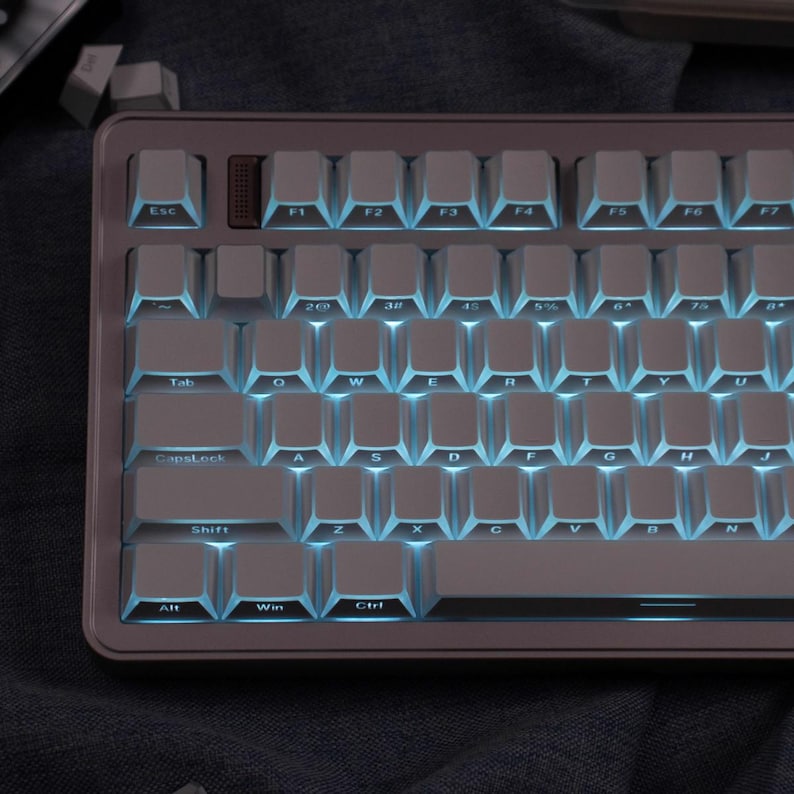 May include: A mechanical keyboard with white keycaps and blue backlighting. The keyboard has a gray frame and a brown keycap in the top row. The keys are labeled with letters, numbers, and symbols. The keyboard is designed for computer use.