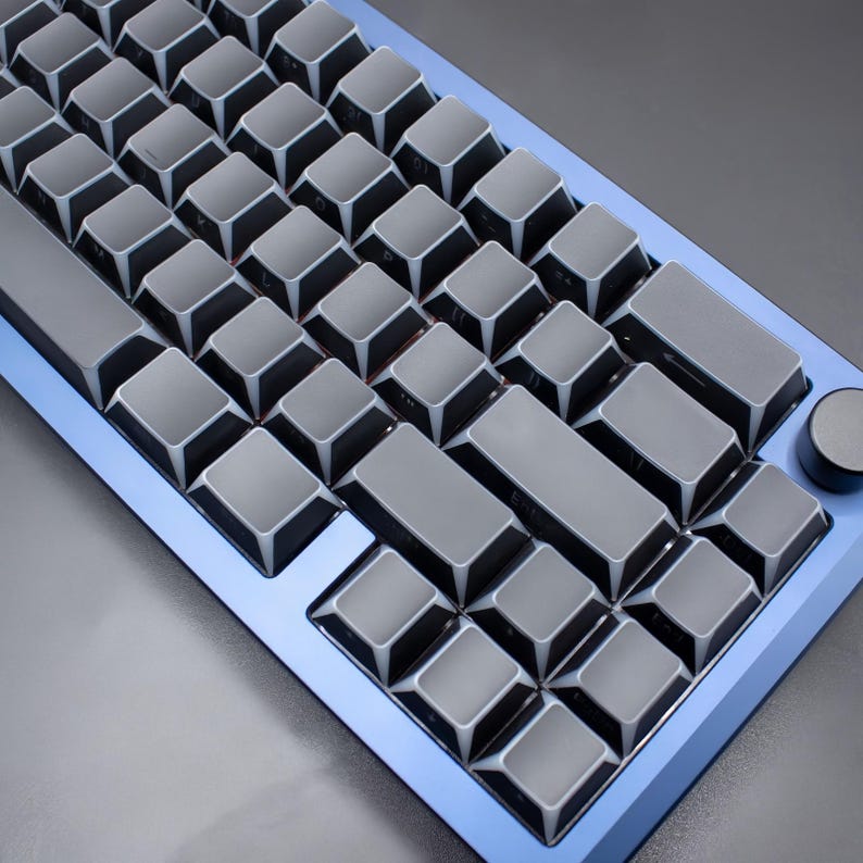 May include: A close-up of a mechanical keyboard with grey keycaps and a blue frame. The keyboard has a compact layout with a volume control knob on the right side. The keycaps are a dark grey colour, and the frame is a vibrant blue.