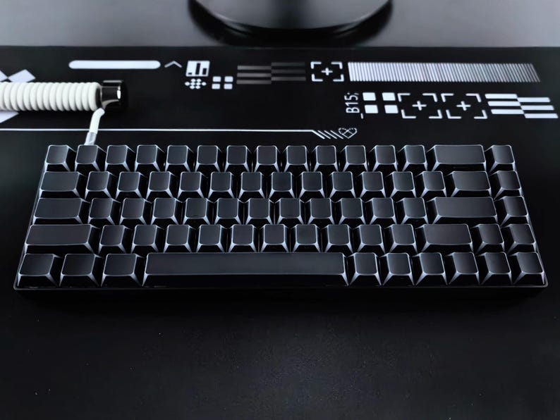 May include: A black mechanical keyboard with square keycaps. The keyboard has a compact layout and a coiled white cable. The background is black with white graphic elements. The keyboard is designed for gaming or typing.