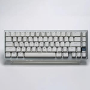 Op de afbeelding: Een zilver-grijze mechanische toetsenbord met witte keycaps. Het toetsenbord heeft een compacte indeling met een transparante plastic cover. De toetsen zijn gemarkeerd met zwarte letters. Het toetsenbord is ontworpen voor computergebruik.