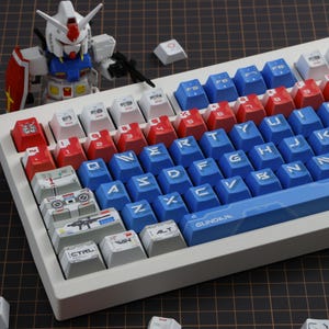 Puede incluir: Un teclado mec&aacute;nico blanco con teclas rojas, blancas y azules. Las teclas presentan varios s&iacute;mbolos y texto, incluyendo "CTRL", "WIN" y "ALT". Una figura de Gundam est&aacute; colocada cerca del teclado.