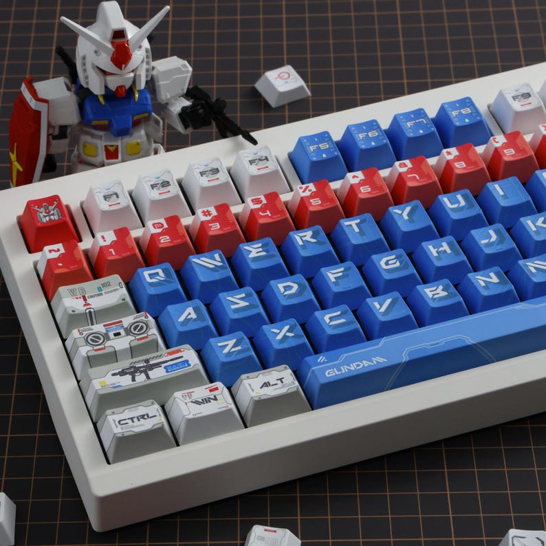Puede incluir: Un teclado mec&aacute;nico blanco con teclas rojas, blancas y azules. Las teclas presentan varios s&iacute;mbolos y texto, incluyendo "CTRL", "WIN" y "ALT". Una figura de Gundam est&aacute; colocada cerca del teclado.