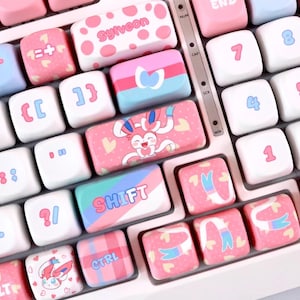 May include: A close-up of a keyboard with custom keycaps in pink, white, and blue. The keycaps feature various designs, including hearts, cartoon characters, and the word "SHIFT". Numbers and letters are also visible.