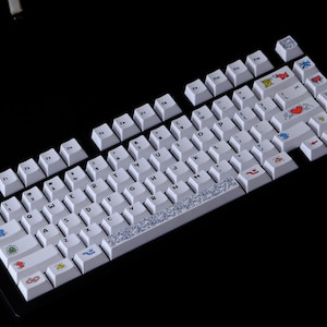 May include: White mechanical keyboard with custom keycaps. The keycaps feature various colorful icons and designs, including hearts, flags, and abstract patterns. The keyboard is set against a black background.