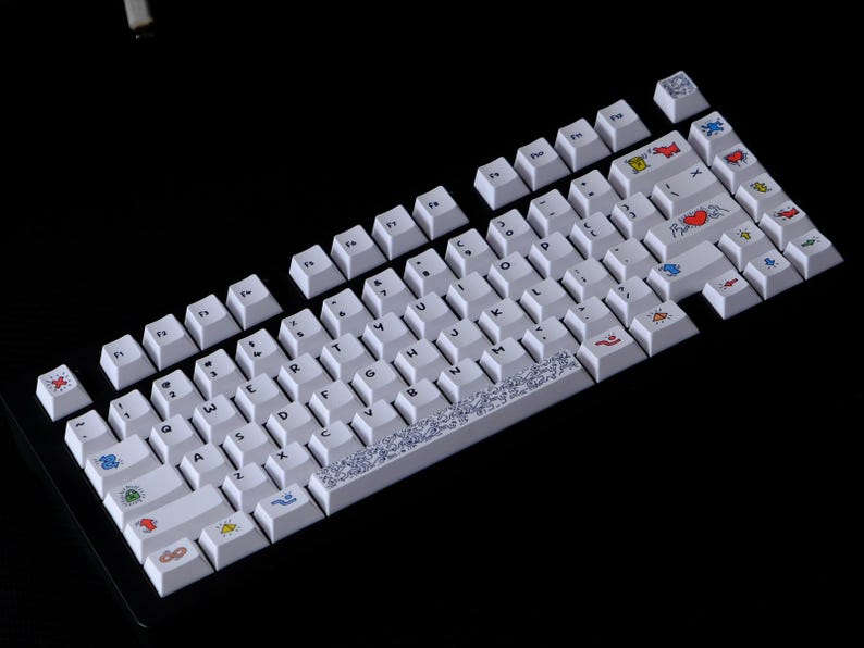 May include: White mechanical keyboard with custom keycaps. The keycaps feature various colorful icons and designs, including hearts, flags, and abstract patterns. The keyboard is set against a black background.